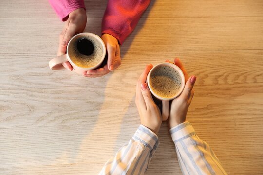 Women with cups of hot coffee at light wooden table, top view