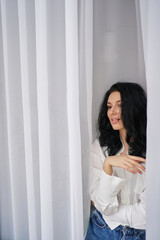 A captivating brunette in jeans and a white shirt strikes a pose amid ethereal white drapes, epitomizing effortless beauty.