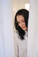 Fototapeta premium A striking brunette in a white shirt exudes timeless charm amid billowing white drapes, a portrait of understated beauty.