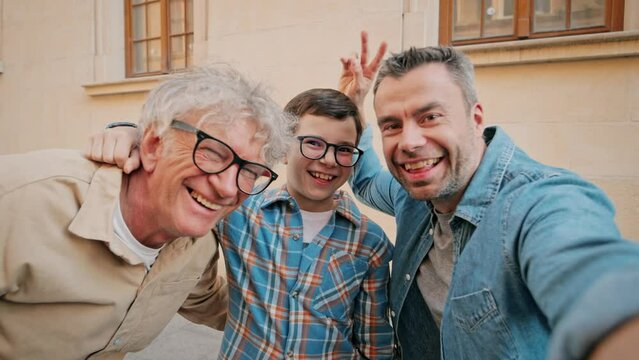Warm moment of three generations having fun together. Caucasian grandson, father and grandfather making funny faces to camera. Dad holding camera for perfect selfie outside in city.