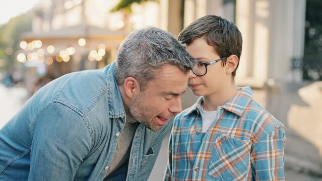 Caucasian middle-aged father leaning down to listen to son in middle of street. Kid whispering something to dads ear. Afterwards man beginning explaining to boy. Child listening with focus.