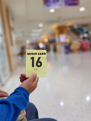 A service user holds a yellow paper queue card with the text Queue Card No. 16 for the X-ray room on blurred background of a hospital room. Patients sit and wait in front of the x-ray to wait service
