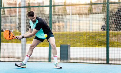 A grown man dressed in sportswear is playing padel on an outdoor court. The boy is hitting the ball...