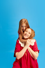 Portrait of beautiful young mother posing with her little daughter in same color dresses against blue studio background. Concept of Mother's Day, International Happiness Day, motherhood, childhood