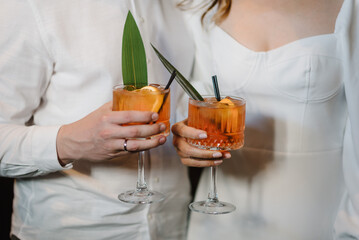 Bride and groom holding cocktails. Man and woman hands hold fresh juice in glass on bar counter at night club. Couple drinking cocktail with slice of orange. Honeymoon concept after wedding. Closeup.