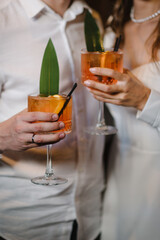 Couple drinking cocktail with slice of orange. Bride and groom holding cocktails. Man and woman hands hold fresh juice in glass on bar counter at night club. Honeymoon concept after wedding. Closeup.