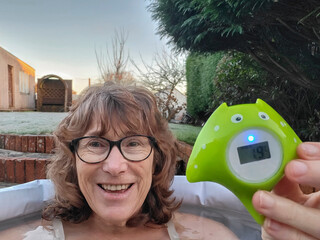 Mature woman takes an ice bath in a pod in her garden. She is holding a thermometer which is reading 1.9 degrees celsius. 