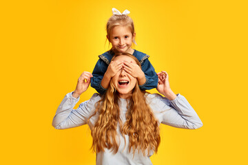 Surprise. Little girl, daughter covering mother's eyes against yellow studio background. Celebrating women's holiday. Concept of Mother's Day, International Happiness Day, family, motherhood - obrazy, fototapety, plakaty