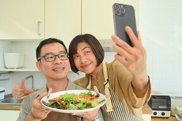 Happy cheerful senior couple taking selfie while preparing health lunch in kitchen.