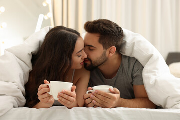 Cute young couple kissing under warm blanket in bed indoors