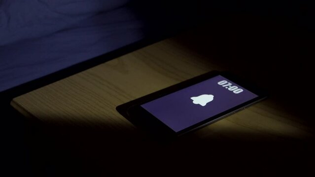 Man wakes up and turns off alarm clock on smartphone. Productive person ready start a day with business opportunities. Close-up focus on mobile phone clock showing six A.M. bedroom apartment
