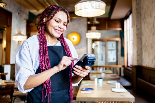 A dark-skinned woman is in uniform working inside a coffee shop. The African girl with colored braided hair is handling a POS terminal. Concept of women working in hospitality. African waitress girls.