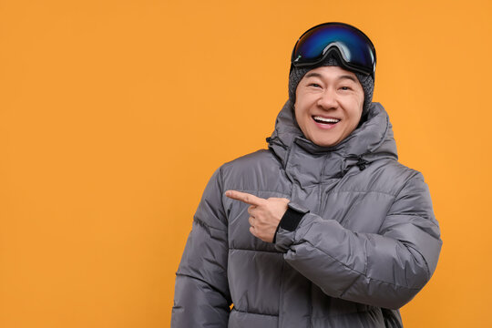 Winter sports. Happy man with ski goggles pointing at something on orange background. Space for text
