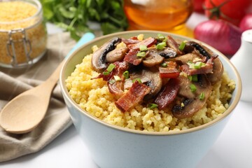 Tasty millet porridge with mushrooms, bacon and green onion in bowl on white table, closeup