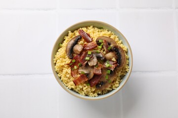 Tasty millet porridge with mushrooms, bacon and green onion in bowl on white tiled table, top view
