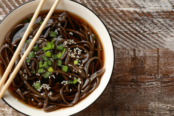 Tasty soup with buckwheat noodles (soba), onion in bowl and chopsticks on wooden table, top view. Space for text