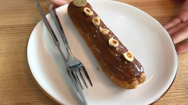 exquisite eclair on white plate with pieces of hazelnut and golden color around knife and fork scroll with hands, looking at delicious dessert, drizzled with milk chocolate. serving yami