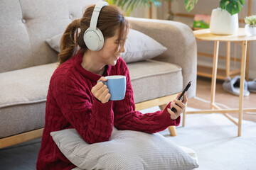 Beautiful young woman in a red casual dress enjoying listening to music and smiling while relaxing on the sofa at home. Young woman with headphones uses laptop and smartphone at home. relax concept.
