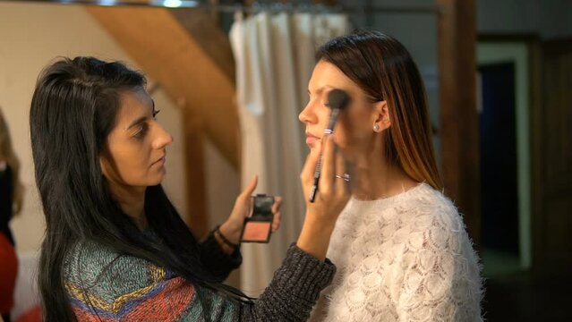 A young actress is doing makeup. The make-up artist applies make-up on the face of a beautiful girl. The model is preparing for shooting in the make-up room. A brunette woman in front of a mirror.
