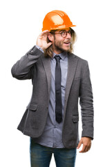 Young handsome architec man with long hair wearing safety helmet over isolated background smiling with hand over ear listening an hearing to rumor or gossip. Deafness concept.