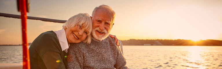 Enjoying amazing sunset. Happy senior couple, elderly man and woman holding hands, hugging and relaxing together while sitting on the side of yacht deck floating in the sea