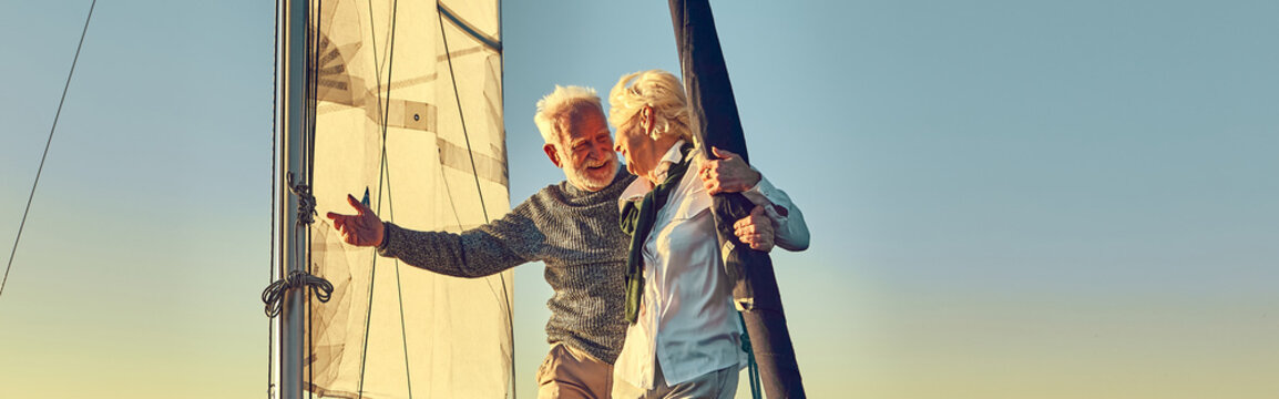 Happy senior man pointing at the horizon while standing with his wife on the side of a sail boat or yacht deck floating in a calm blue sea
