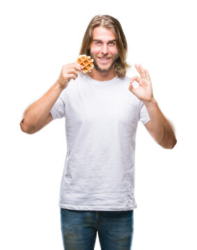 Young handsome man with long hair over isolated background eating waffle doing ok sign with fingers, excellent symbol