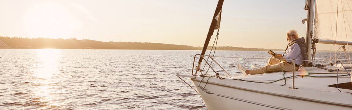 View of the sail boat or yacht floating in sea with relaxed senior man reading a book, sitting on the deck