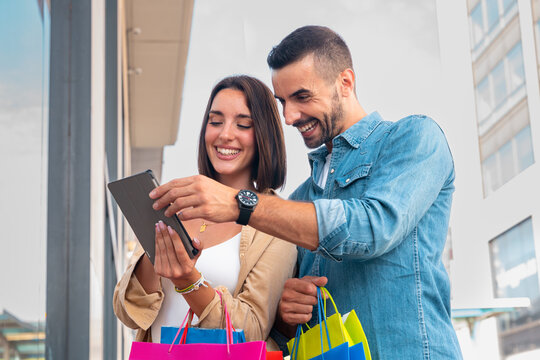 Smiling young Caucasian couple holding shopping bags, browsing tablet apps