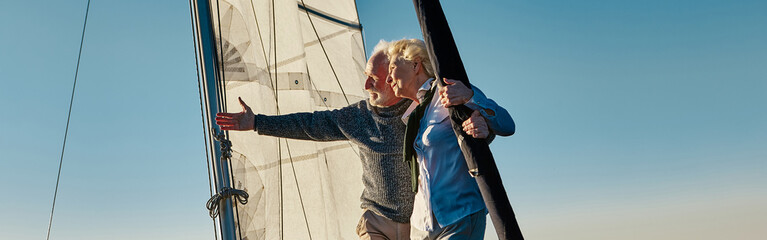 Elegant senior man pointing at the horizon while standing with his wife on the side of a sail boat...