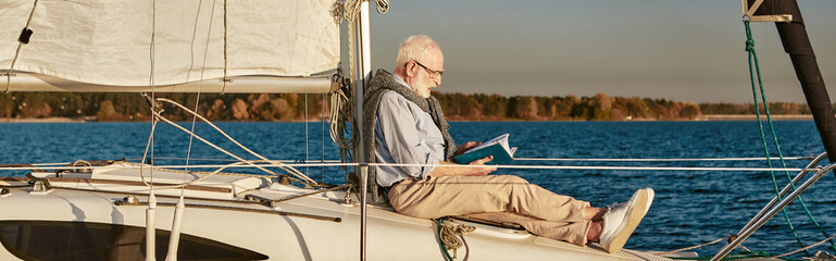 Senior man reading book and relaxing while sitting on the side of sailboat or yacht deck floating...