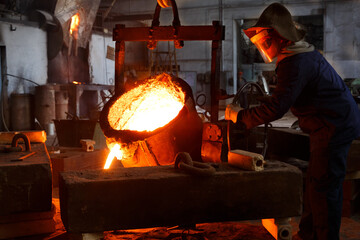 Industrial worker in protective gear pours molten metal at steel foundry. Manufacturing process in...