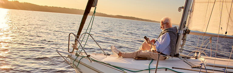 Reading on board. Side view of a relaxed senior man sitting on the side of sailboat or yacht deck...