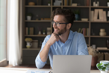 Pensive smiling millennial business man in glasses distracted from computer work look in distance...