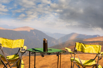 Tourist chairs and a table with a thermos in a clearing overlooking the mountain peaks on a sunny...