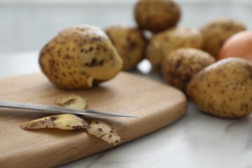 Fresh potatoes, peels, wooden board and knife on white marble table, closeup