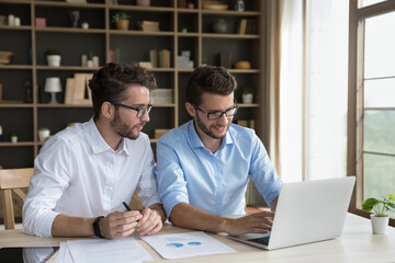Two male twins sit at desk using laptop, millennial 35s brothers having business partnership, joint business, working on project, prepare marketing strategy, busy in startup, workflow in home-office - obrazy, fototapety, plakaty