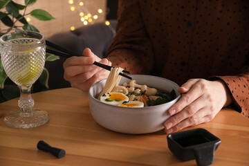 Woman eating delicious ramen with chopsticks at wooden table indoors, closeup. Noodle soup