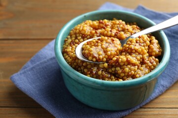 Whole grain mustard in bowl and spoon on wooden table, closeup