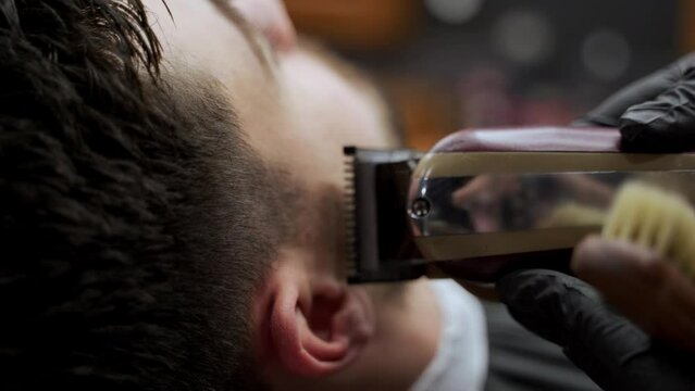 Barber shapes clients hairstyle with electric clipper in barbershop. Professional hairstylist trims side of mans head. Grooming session, precise haircut for stylish male. Expert barber at work.