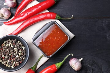 Spicy chili sauce, garlic, peppers and peppercorns on black wooden table, top view. Space for text