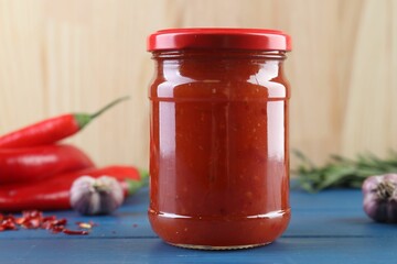 Spicy chili sauce in jar, garlic and peppers on blue wooden table, closeup