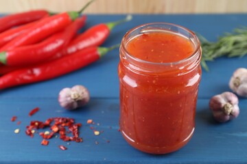 Spicy chili sauce in jar, garlic, peppers and rosemary on blue wooden table, closeup