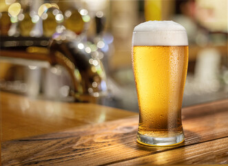 Glass of chilled beer on wooden bar table top and blurred bar interior at the background.