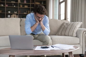 Stressed single man feels frustrated due to overspend, high household rates, unpaid taxes, lack of finances to pay domestic bills sit at table with laptop and heap of papers. Penalty, eviction, crisis - obrazy, fototapety, plakaty