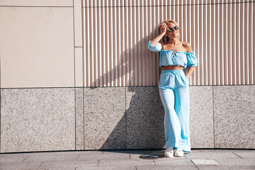 Portrait of young beautiful smiling hipster woman in trendy summer clothes. Sexy carefree woman posing in the street at sunset. Positive blond model outdoors. Cheerful and happy. Near wall