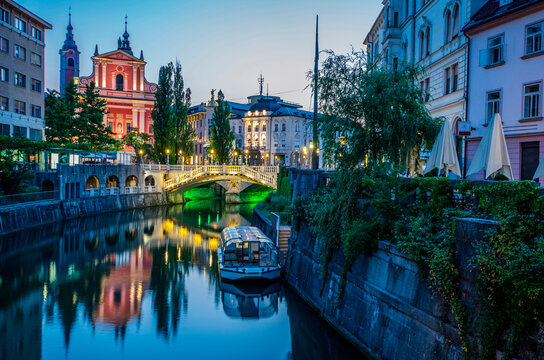 Fototapeta Twilight view of a calm river passing through Ljubljana, with illuminated historical buildings and a bridge reflecting on water