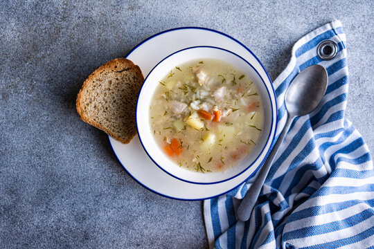 A bowl of vegetable and chicken soup accompanied by a slice of bread placed on a gray surface with a blue and white striped napkin and a spoon to the side