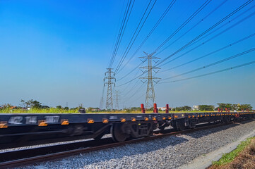 Fototapeta premium a series of flat-car trains speeding by against the backdrop of high-voltage power lines during a clear blue sky