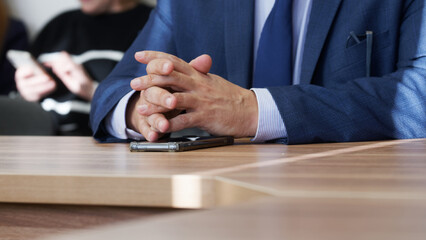 Folded hands of a politician, official or businessman on a table with smartphone. Working meeting with colleagues or negotiations. Manager or lawyer. Without face. Photo. Selective focus - obrazy, fototapety, plakaty